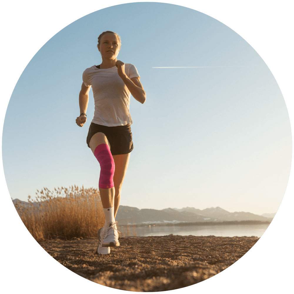 Woman wearing Bauerfeind's Sports Compression Knee Support while running on a grassy trail near a lake.