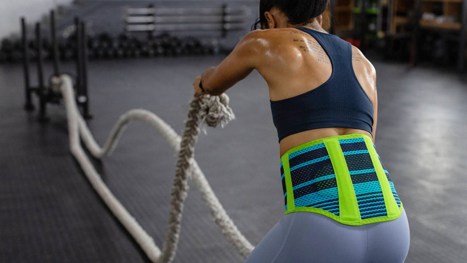 Woman wearing Bauerfeind's Sports Back Support while performing a rope exercise in an indoor gym.