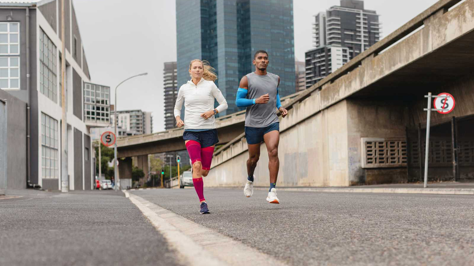 Two young adults running on an inner-city street while wearing Bauerfeind's Sports Compression Sleeves for the arm, upper leg and lower leg.