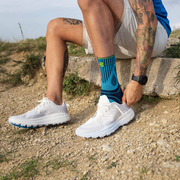 Man donning his shoes while wearing Bauerfeind's Sports Achilles Support before going on a morning jog through a grassy field.