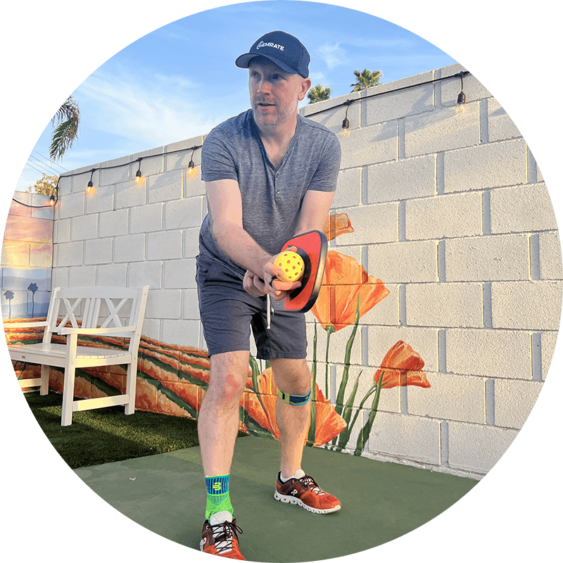 Man preparing a pickleball serve while wearing Bauerfeind's Sports Ankle Support and Sports Knee Strap.