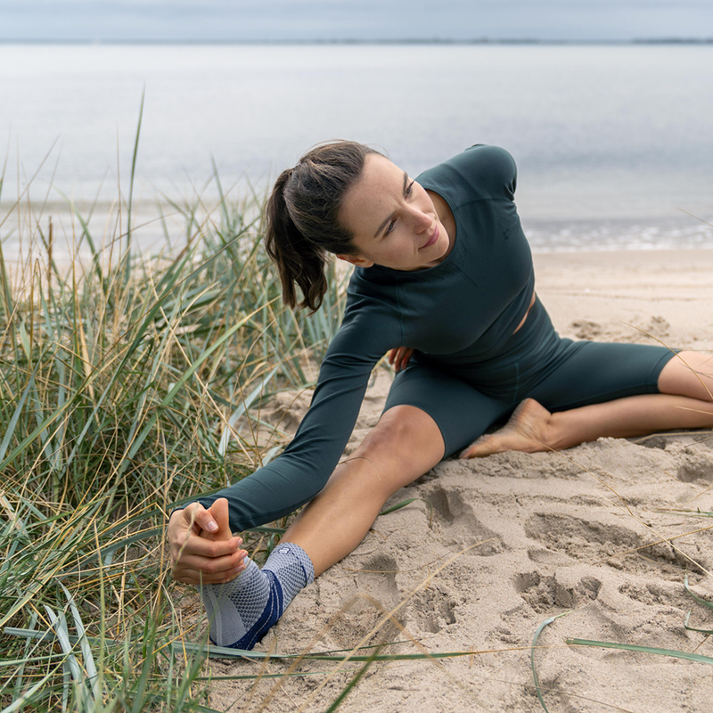 Woman practicing yoga on a sandy beach overlooking a serene lake, wearing a green workout outfit and a supportive ankle brace.