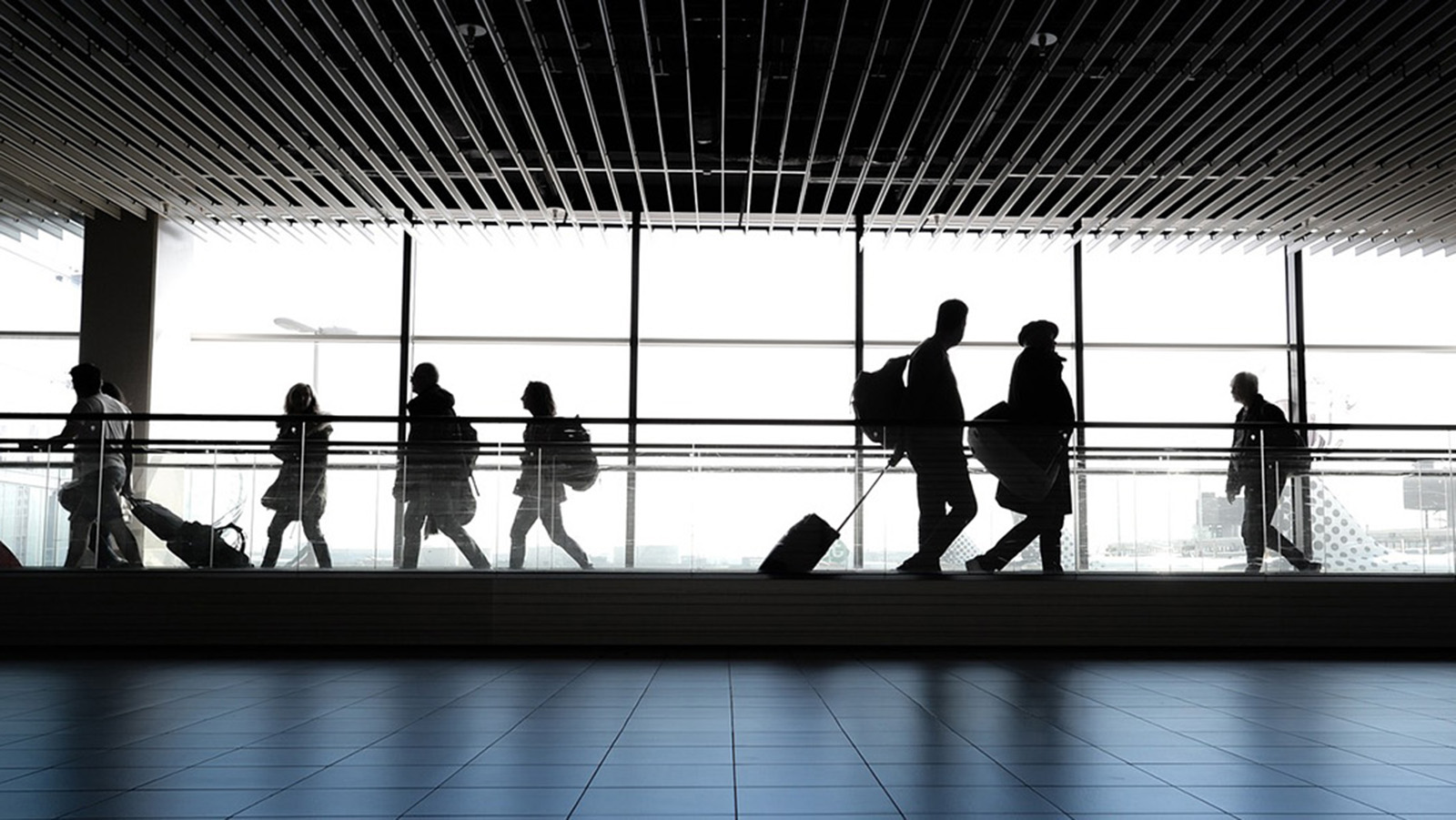 Silhouettes of people walking in an airport terminal with large windows and modern architecture, highlighting the hustle of travel.