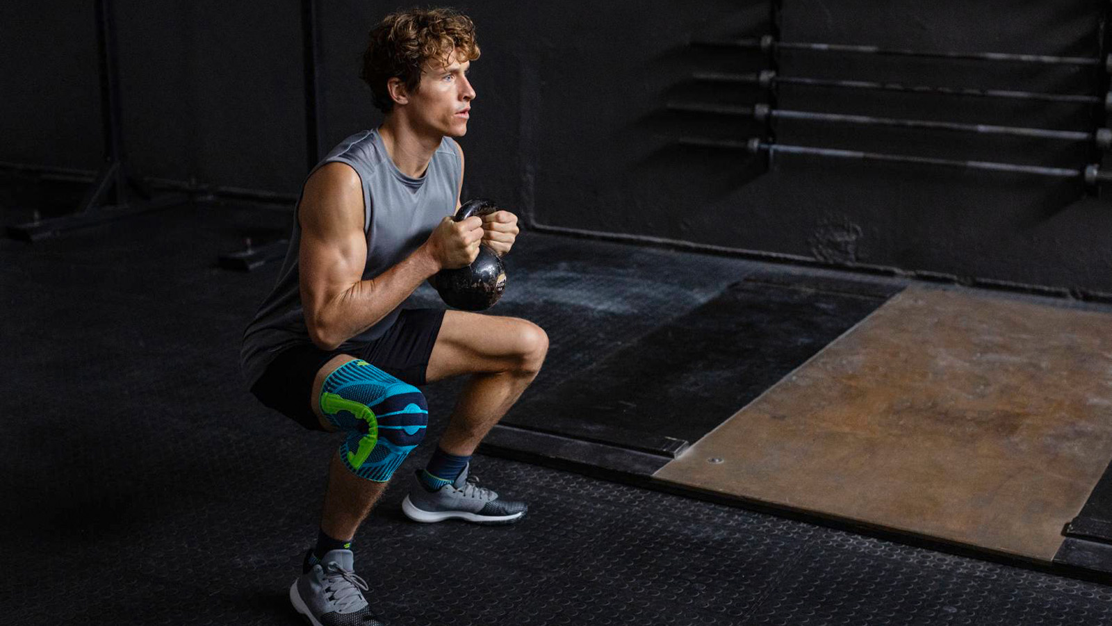 Man wearing Bauerfeind's Sports Knee Support inside of a fitness center while performing kettlebell squats.