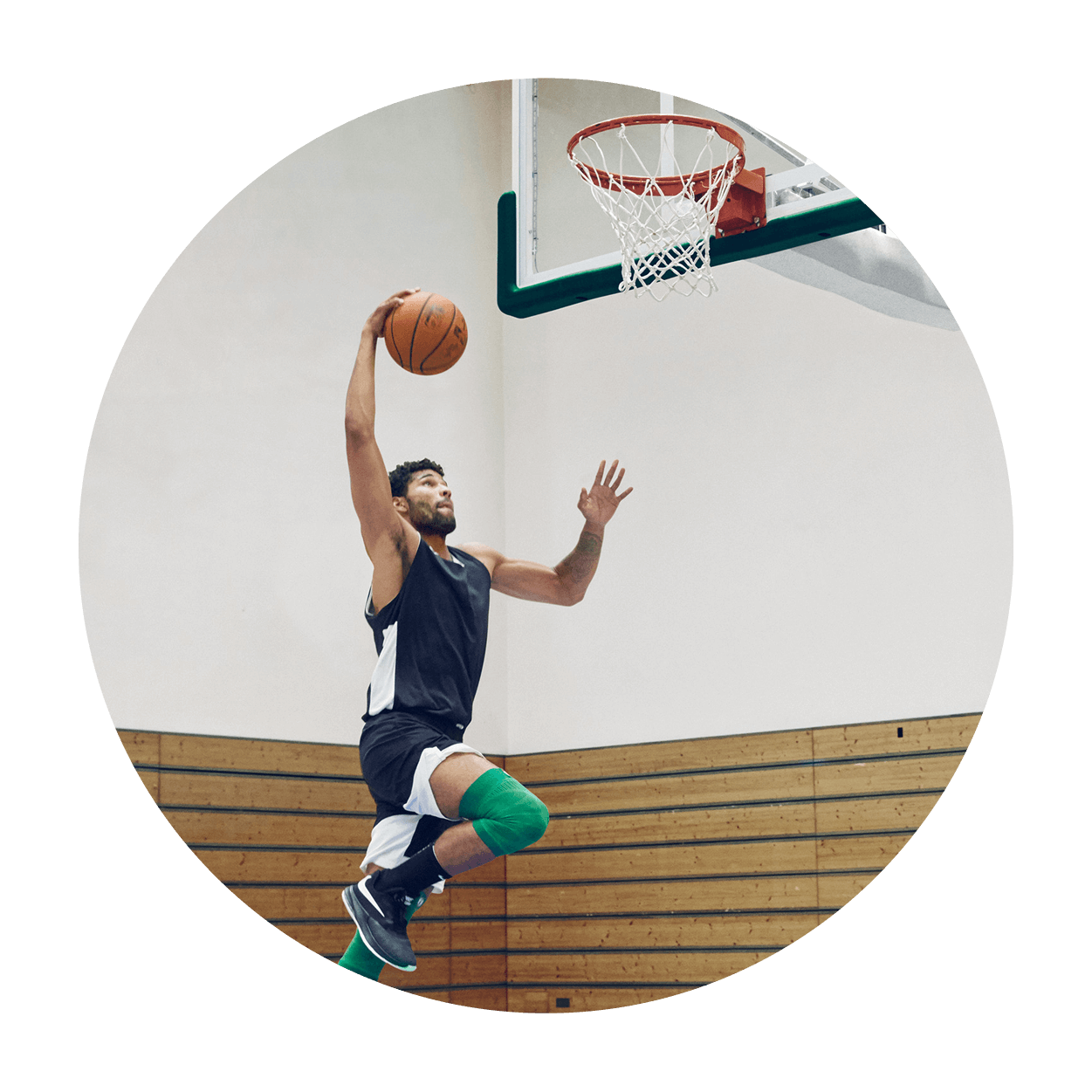 Athlete attempting a dunk on an indoor basketball court while wearing Bauerfeind's Sports Compression Knee Support NBA Celtics Edition.