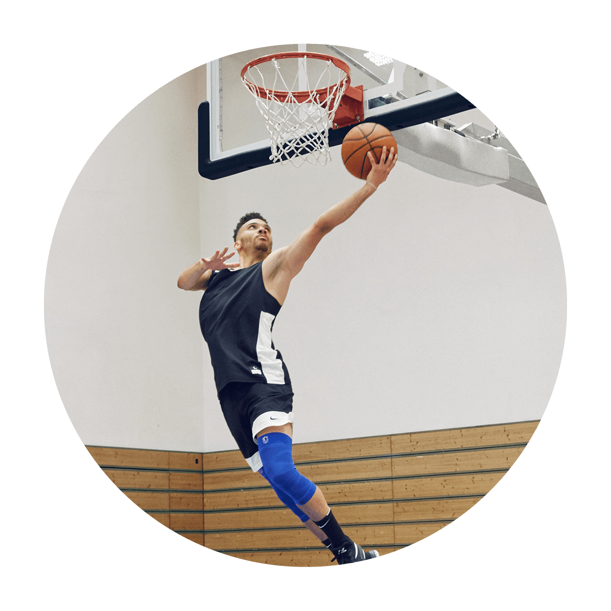 Athlete attempting a layup on an indoor basketball court while wearing Bauerfeind's Sports Compression Knee Support NBA Mavericks Edition.