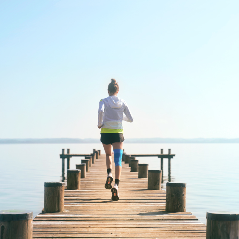 Woman wearing Bauerfeind's Sports Knee Support while running through a forest trail.