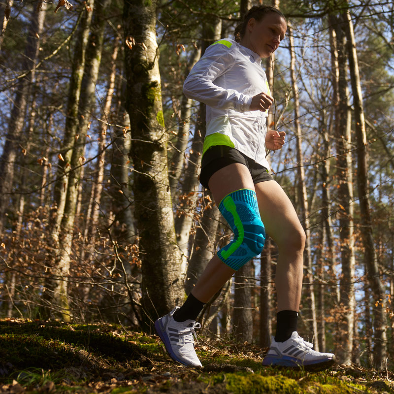 Woman wearing Bauerfeind's Sports Knee Support while running through a forest trail.