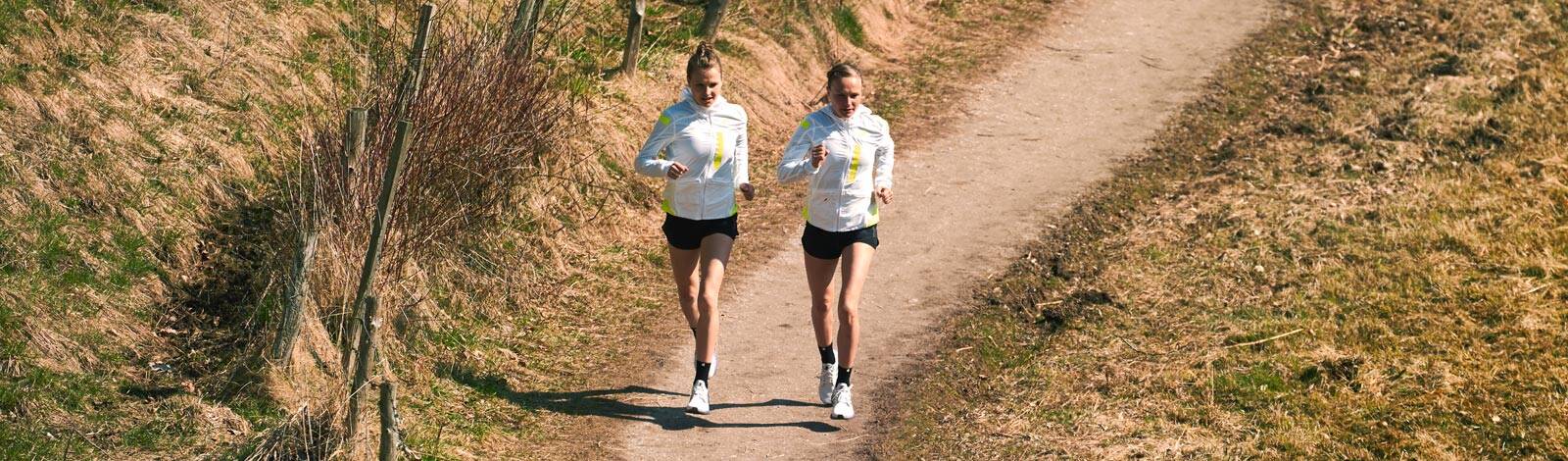 Banner image of two women jogging on a dirt path in a rural setting, dressed in white and yellow athletic wear.