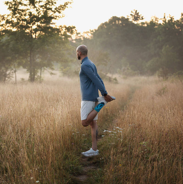 Man wearing Bauerfeind's Sports Achilles Support while stretching before a morning run through a grassy field.