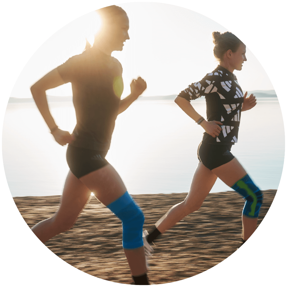 Two women running near a lake, with the runner on the left wearing Bauerfeind's Sports Compression Knee Support and the runner on the right wearing the Sports Knee Support.