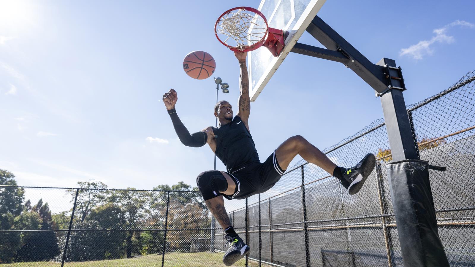 Athlete wearing Bauerfeind's Sports Knee Support NBA while dunking on an outdoor basketball court.