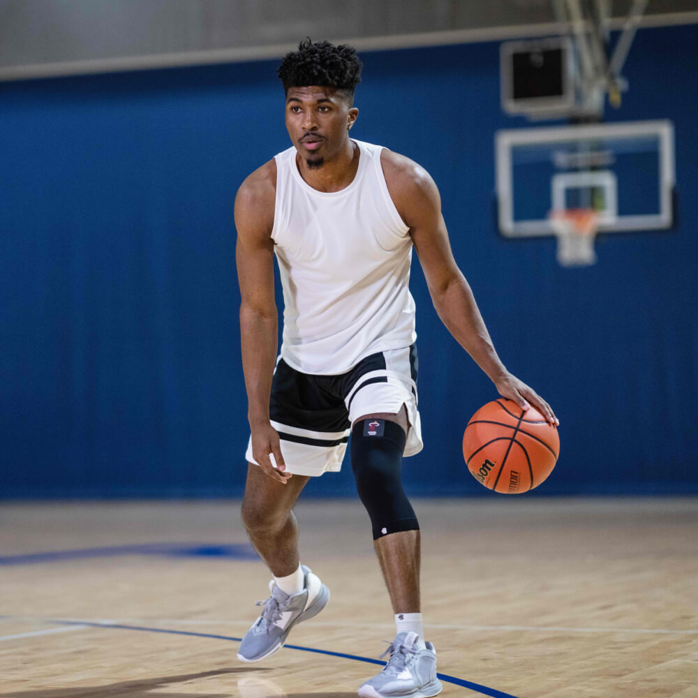 Basketball player dribbling a basketball on an indoor court wearing white tank top and black shorts.