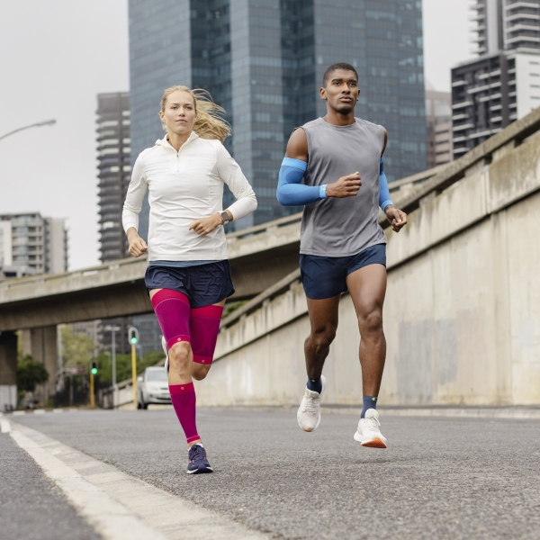 Two young adults running on an inner-city street while wearing Bauerfeind's Sports Compression Sleeves for the arm, upper leg and lower leg.