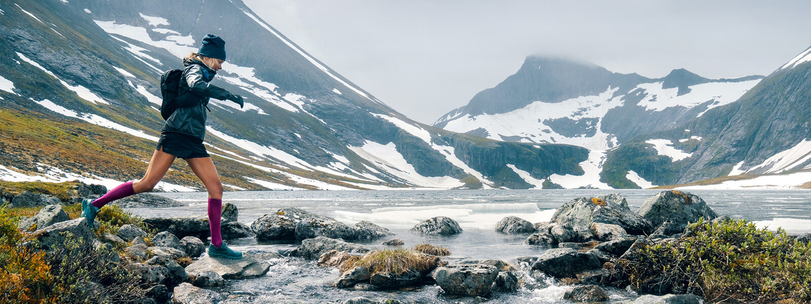 Image of woman running across a creek wearing Bauerfeind's Run Ultralight Compression Socks with a mountainous landscape in the background.