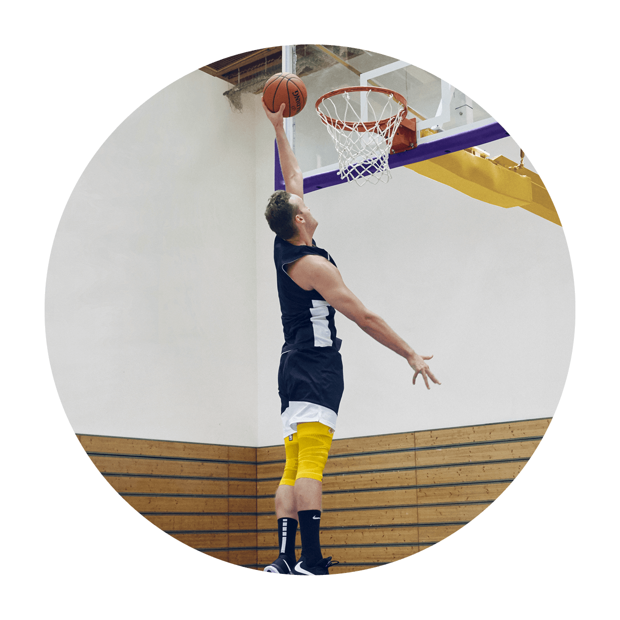 Athlete attempting a dunk on an indoor basketball court while wearing Bauerfeind's Sports Compression Knee Support NBA Lakers Edition.