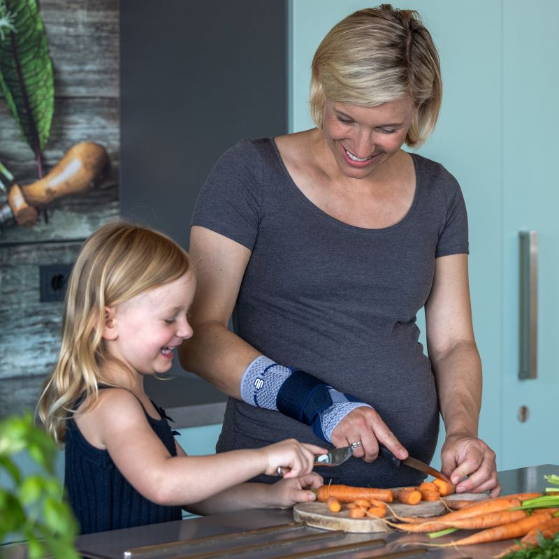 Image of a woman wearing Bauerfeind's ManuTrain wrist brace (titan) while cutting carrots in the kitchen with her daughter.