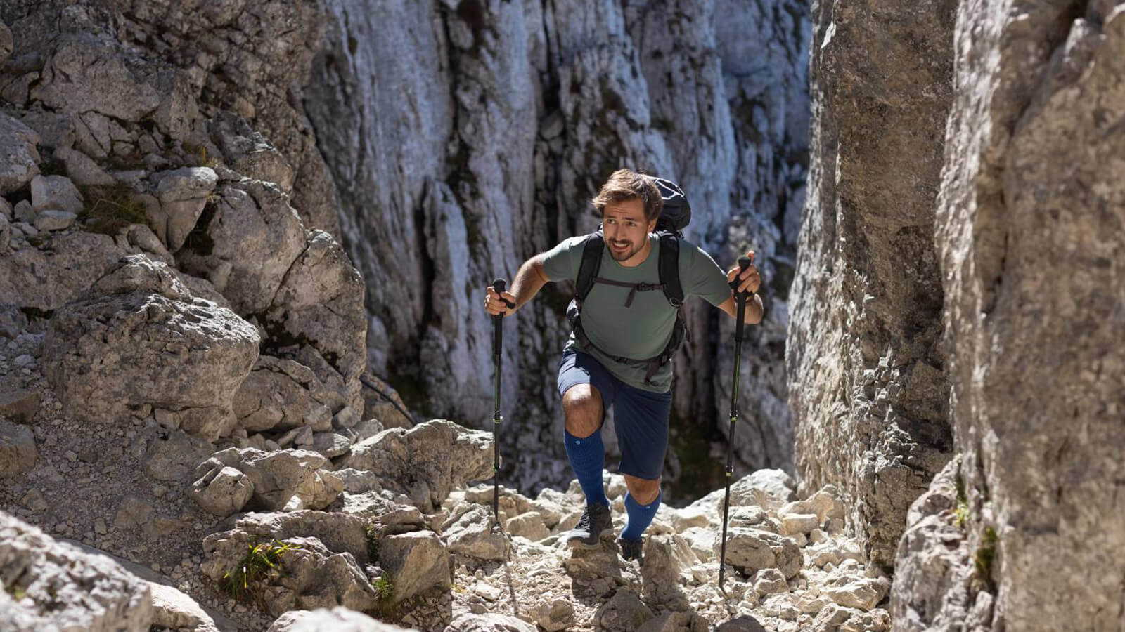 Man wearing Bauerfeind's Outdoor Merino Compression Socks while hiking through rocky mountain terrain using trekking poles.