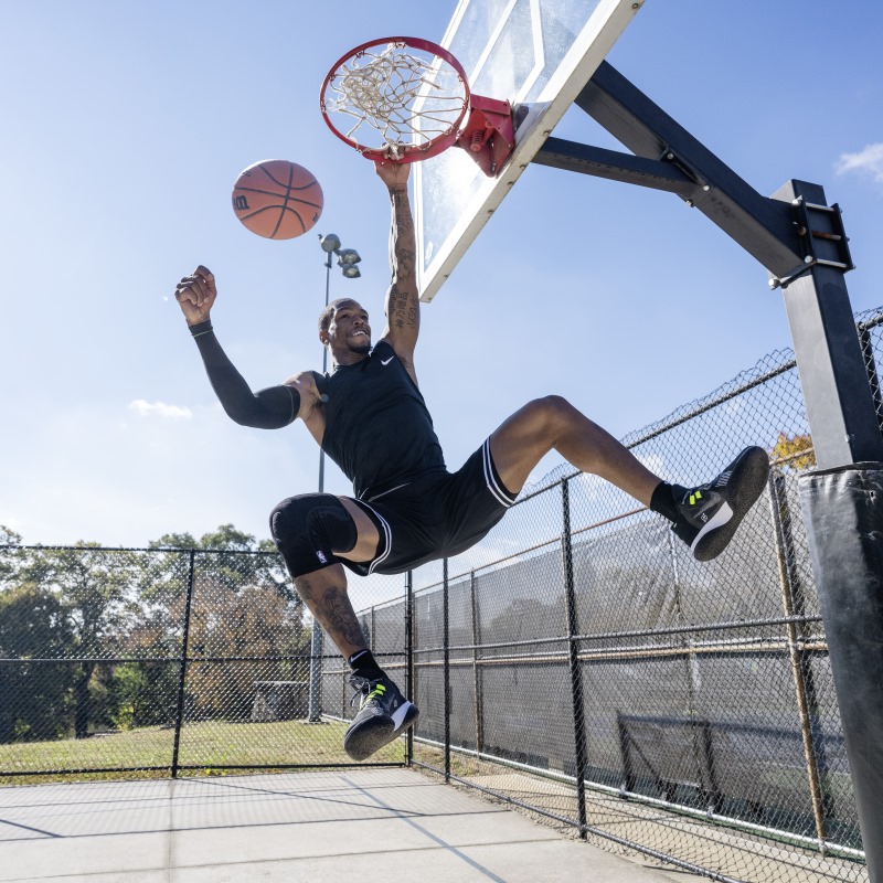 Athlete wearing Bauerfeind's Sports Knee Support NBA while dunking on an outdoor basketball court.