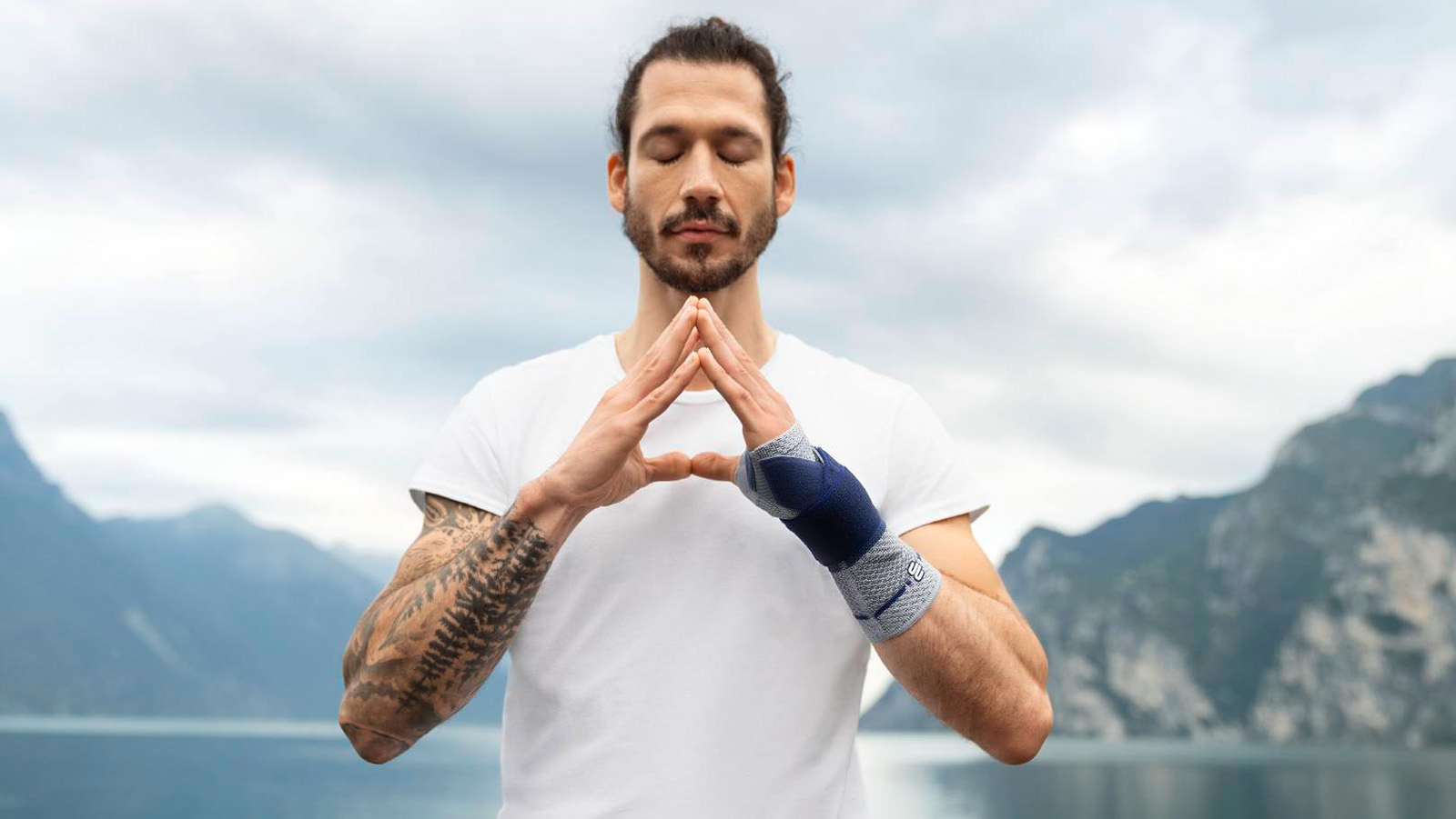 Man wearing Bauerfeind's ManuTrain wrist brace while practicing yoga and mindfulness meditation outdoors near a lake and mountains.