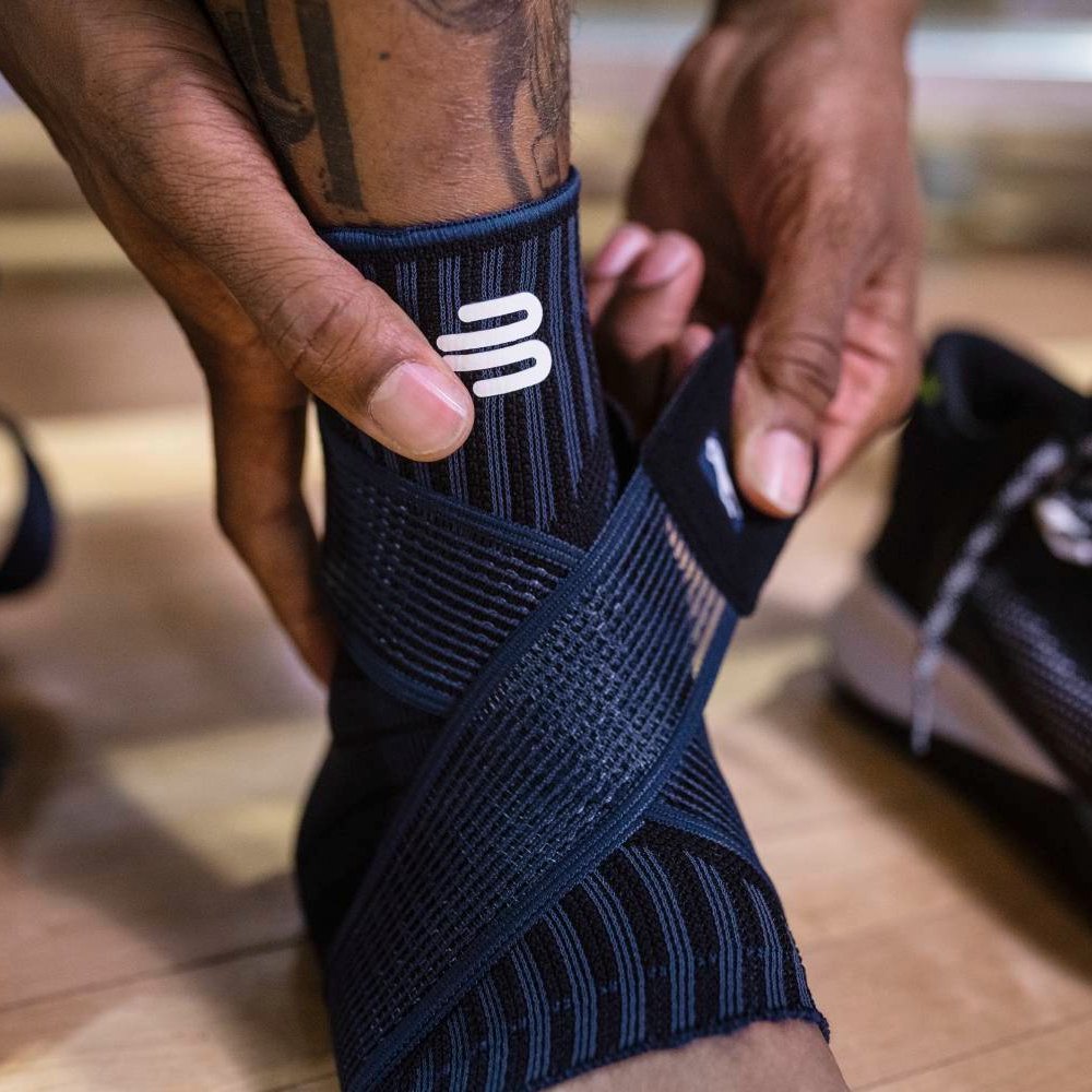 Close-up of hands adjusting a black compression ankle brace on a wooden floor.