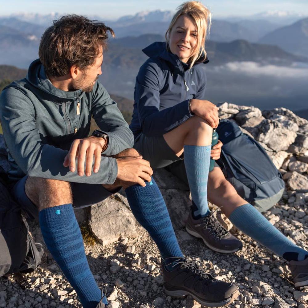 Hikers enjoying a mountain view while resting on rocky terrain, wearing blue and green outdoor gear and knee-high compression socks.