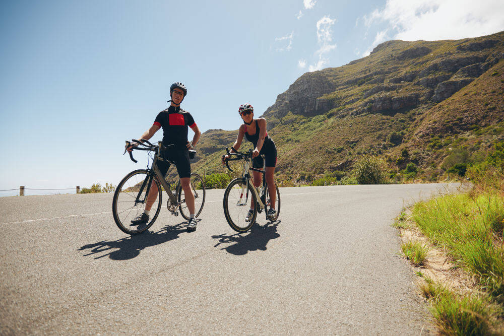 Two cyclists in sportswear standing next to their road bikes on a sunny mountain road.