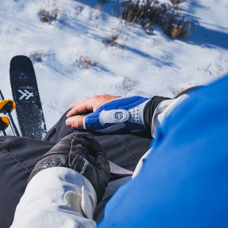 Image of a skier wearing Bauerfeind's RhizoLoc thumb spica splint while sitting on a chairlift above a snowy mountain.