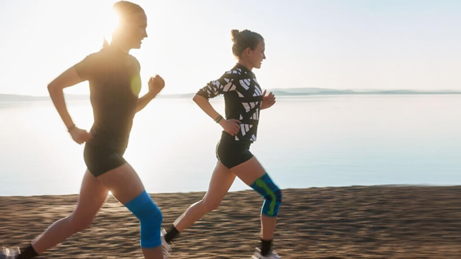 Two women running on the sand near the ocean. The runner on the left is wearing Bauerfeind's Sports Compression Knee Support, and the runner on the right is wearing Bauerfeind's Sports Knee Support.