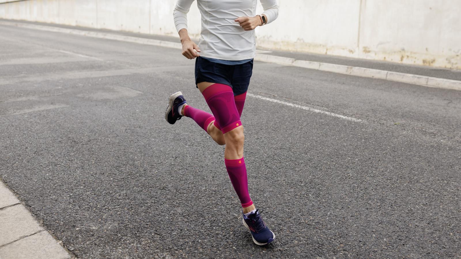 Image of a woman wearing Bauerfeind's Sports Compression Upper Leg Sleeves and Sports Compression Lower Leg Sleeves while running through a city street.