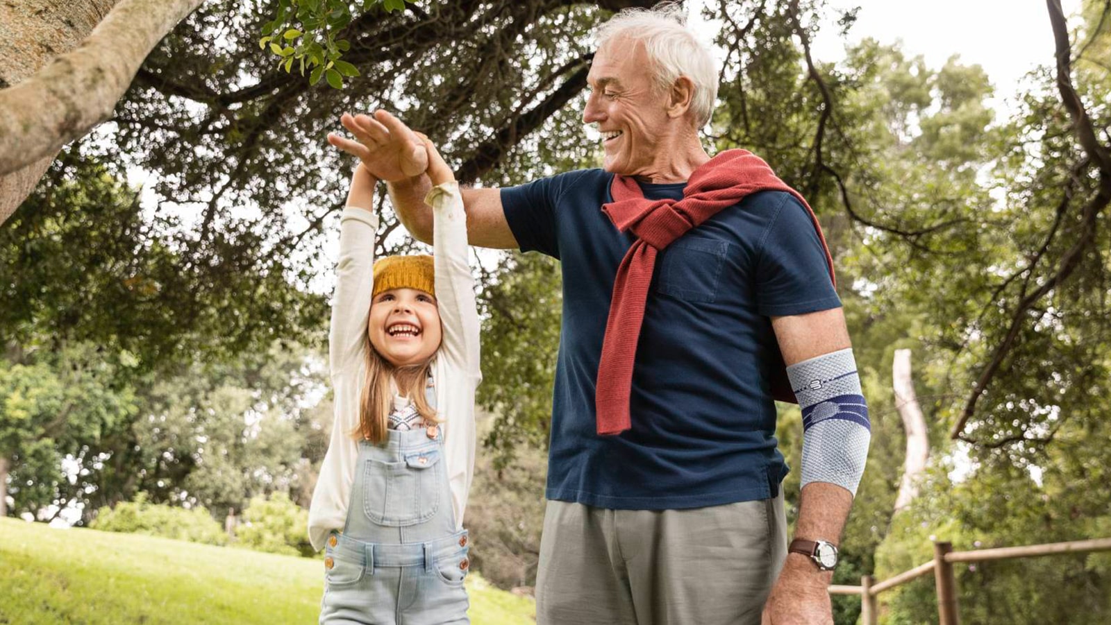 Image of a Grandfather wearing Bauerfeind's EpiTrain elbow brace while spending time with Granddaughter in a park.