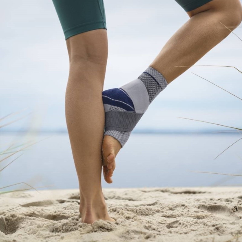 Close-up view of a person stretching on a sandy beach, wearing a grey and blue knee support brace and teal athletic shorts, with a calm lake and sky in the background.