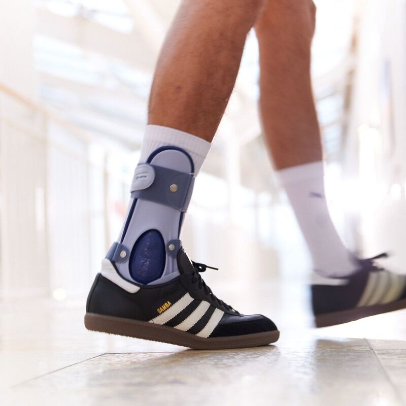 Close-up of a person walking with an orthopedic ankle brace and black Adidas Samba sneakers.