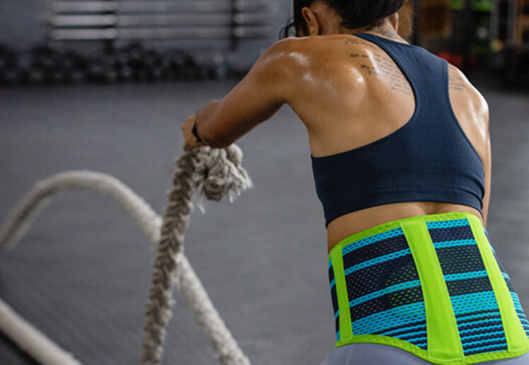 Woman wearing Bauerfeind's Sports Back Support while performing a rope exercise in an indoor gym.