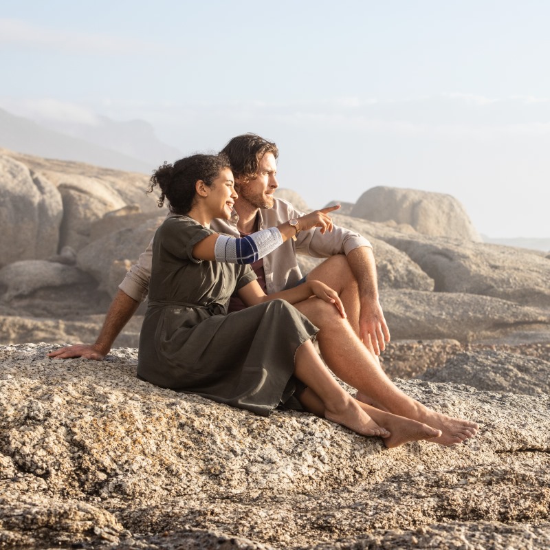 Couple sitting on rocky coast and pointing towards ocean, with the woman wearing Bauerfeind's EpiTrain elbow brace.