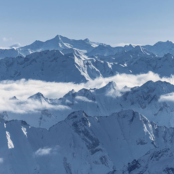 Aerial view of majestic snowy mountain peaks with clouds hovering around, in bright daylight.