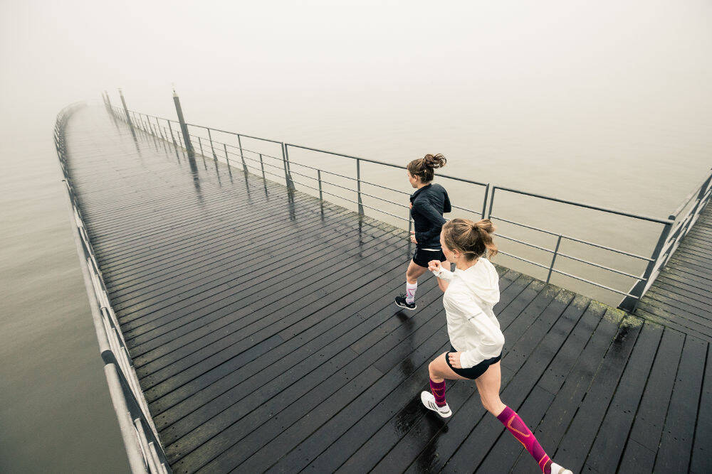 Two women wearing Bauerfeind's Run Performance Compression Socks while running on a foggy wooden pier stretching into a lake.