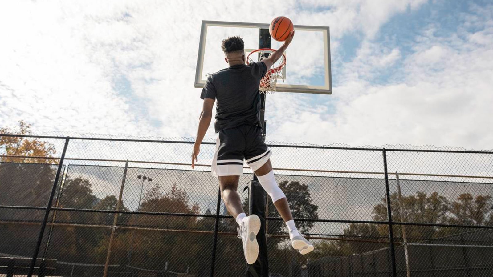 Basketball player wearing Bauerfeind's Sports Compression Knee Support NBA while attempting a layup at an outdoor court.