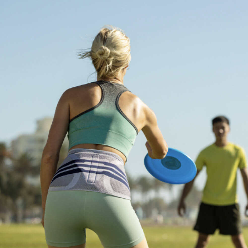 Woman wearing Bauerfeind's LumboTrain waisted back brace while throwing a frisbee to a man in the park.