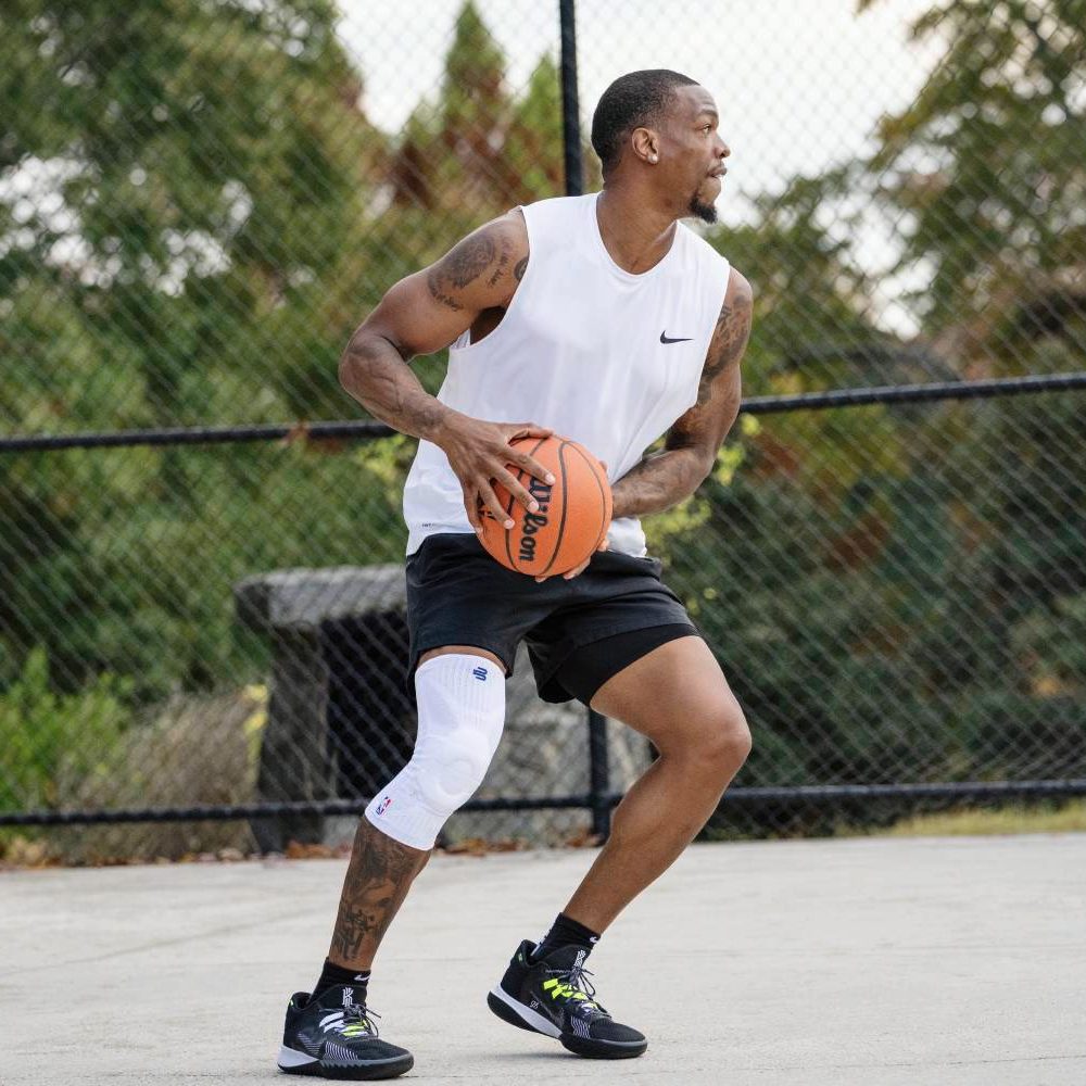 Man playing basketball outdoors wearing white tank top and knee support.