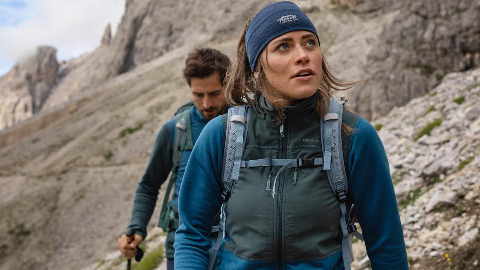 Young man and woman hiking in rugged mountain terrain, both wearing backpacks and outdoor gear, focused on the trail ahead.