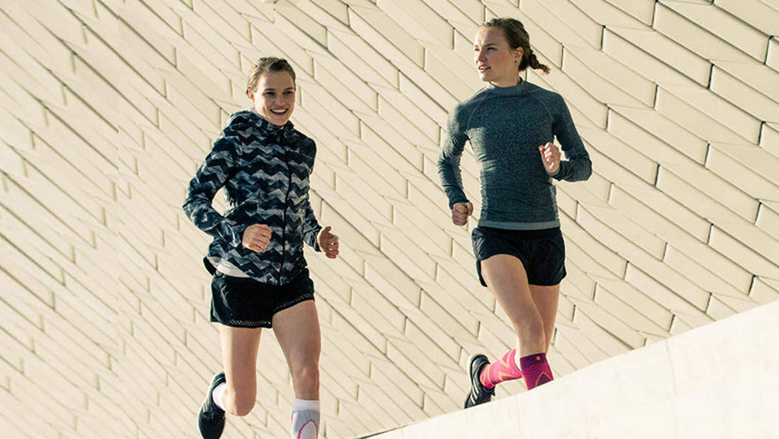 Two young women wearing Bauerfeind's Run Performance Compression Socks while running on a concrete path inside of a city.