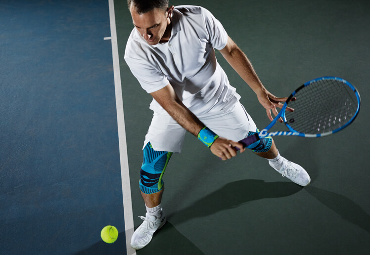 Tennis player wearing Bauerfeind's Sports Knee Support and Sports Wrist Strap while attempting to hit the tennis ball.