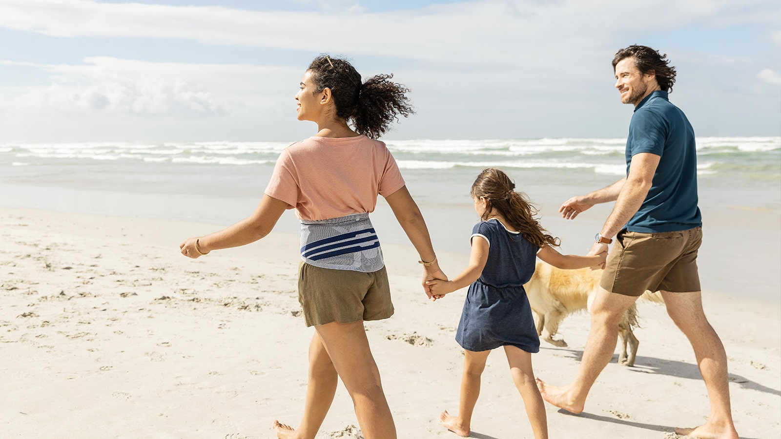 Family walking on the beach with their dog, while the mother wears Bauerfeind's LumboTrain waisted back brace.