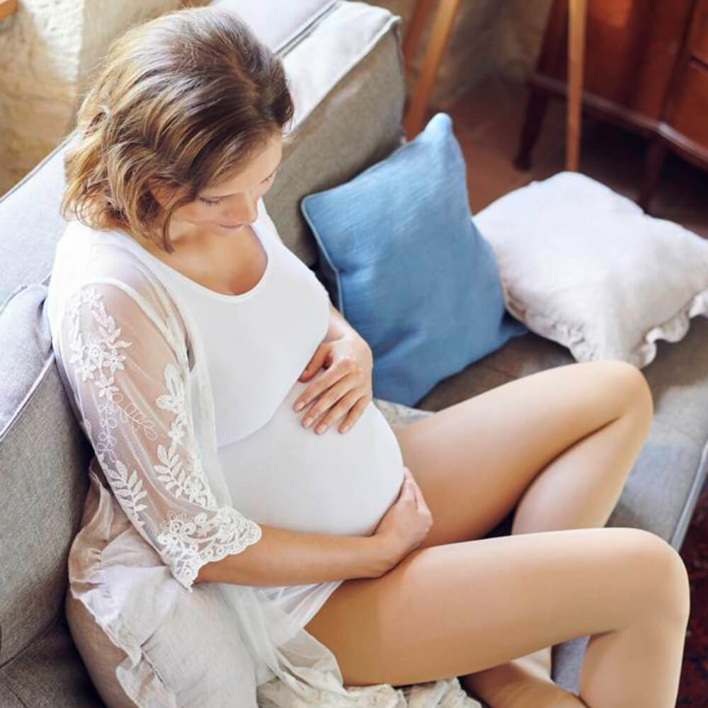 Pregnant woman in white dress sitting on a sofa and wrapping her arms around her belly while wearing Bauerfeind's VenoTrain micro Compression Stockings.