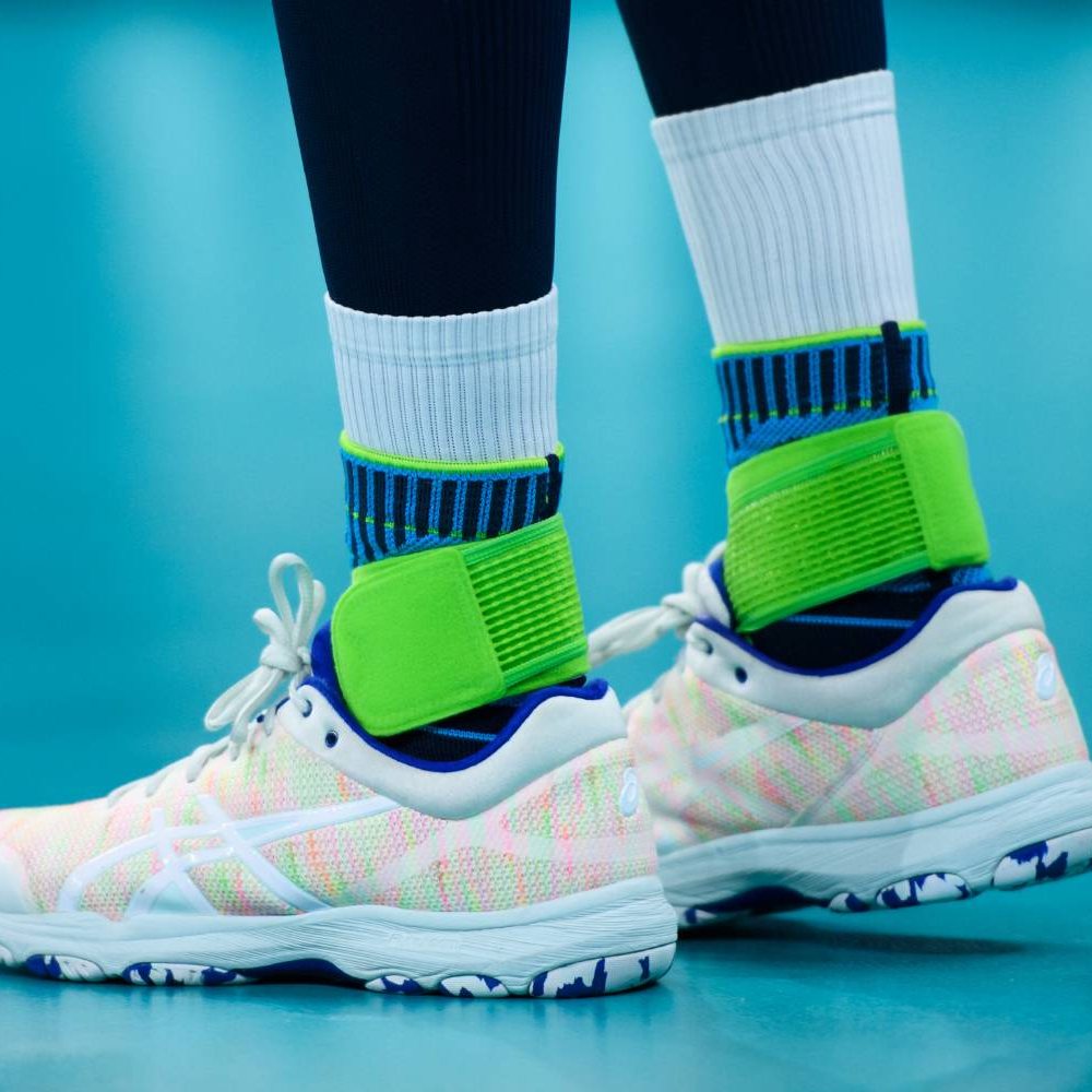 Close-up of a person wearing white sport shoes with neon green ankle supports, on blue indoor court.