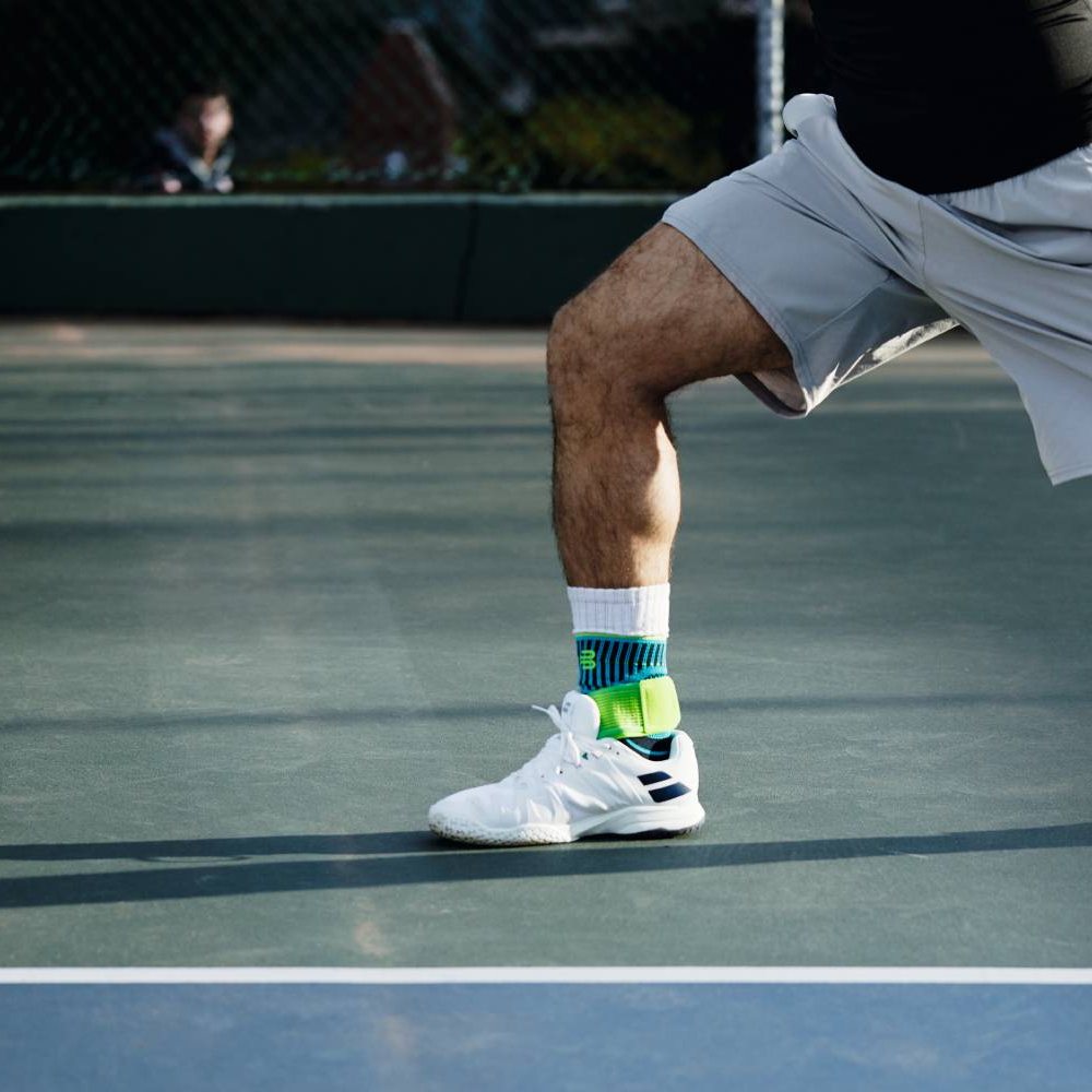 Close-up of a person wearing white sport shoes with neon green ankle supports, on blue indoor court.