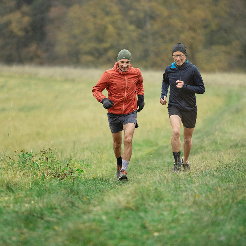 Two adult men jogging in a field during fall, one wearing a red jacket and one in a blue jacket, both in exercise gear on a cloudy day.