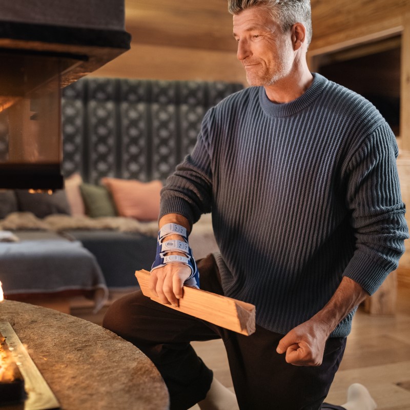 Image of a man wearing Bauerfeind's ManuLoc Rhizo wrist and thumb brace while placing wood onto a chimney fire.