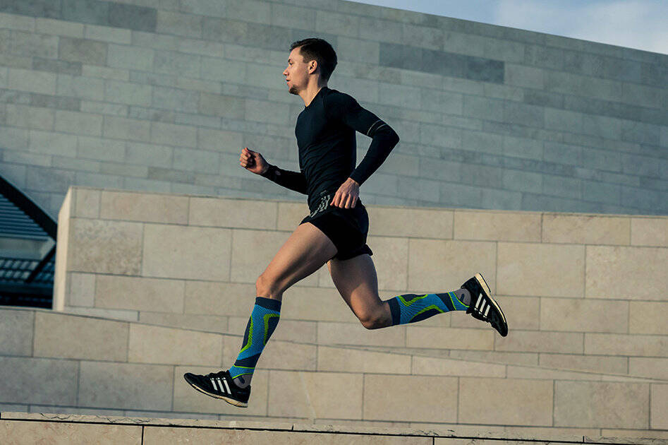 Young man wearing Bauerfeind's Run Performance Compression Socks while running on a concrete path inside of a city.